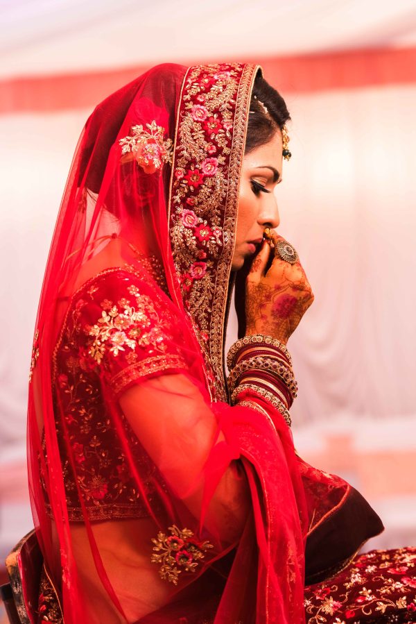 The bride in a red saree is sitting with her hands folded